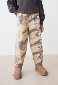 Pantalon camouflage beige et gris avec une ceinture élastique, associé à des bottines marron clair en tissu doux.
