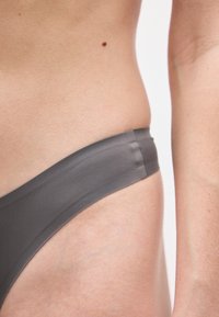 Κοντινό πλάνο της μέσης και του χεριού ενός ατόμου, που δείχνει λείο δέρμα, μια μικρή ελιά και τη ζώνη της γκρίζας εσωτερικής παντόφλας.