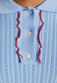 Light blue short-sleeve polo shirt featuring a ribbed texture, subtle vertical stripes, red ruffled trim along the collar, and white buttons.