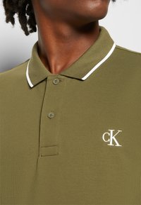 Polo vert olive avec un tissu texturé, un col blanc, une patte de boutonnage à deux boutons et un petit logo "CK" brodé sur la poitrine.