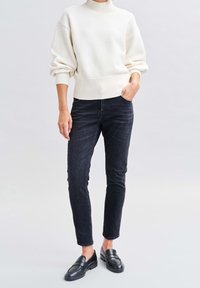 Osoba v krémovém žebrovaném svetru, černých slim fit džínách a černých mokasínech, s jednou rukou v kapse, stojící na jednobarevném pozadí.