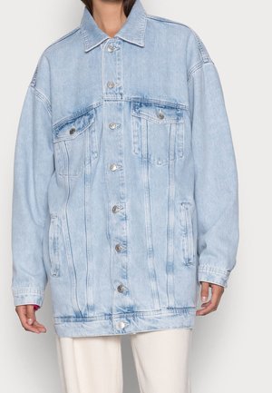 Personne portant une veste en jean bleu clair oversize avec fermetures à boutons, poches poitrine à rabat et poches latérales fendues, associée à un pantalon clair.