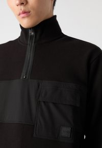 Felpa nera con zip e colletto alto, caratterizzata da una sezione superiore testurizzata e una tasca frontale piatta accentuata da un logo.