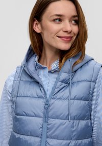 Hellblau gepolsterte Weste mit Kapuze, die vertikale Nähte und einen Reißverschluss vorne hat. Darunter ist ein blau-weiß gestreiftes Hemd mit Kragen sichtbar.