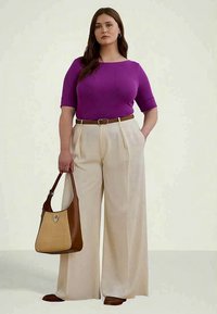 Mulher de pé, vestindo uma blusa roxa, calças largas bejes com um cinto castanho, segurando uma bolsa castanha e bege, em um fundo liso.