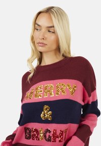 Maglione a righe bordeaux, rosa e blu navy con la scritta "MERRY & BRIGHT" in lettere glitterate dorate e rosse; tessuto morbido e vestibilità comoda.