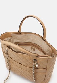 Beżowa torba tote pikowana z dwoma uchwytami, łańcuszkowym paskiem, przednim logo "GUESS" oraz otwartym górnym zamknięciem, które eksponuje wnętrze z kieszeniami na zamek i podszewką materiałową.