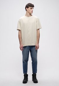 Smėlio spalvos medvilniniai marškinėliai su trumpomis rankovėmis ir moderniu kirpimu, derinami su mėlynais siaurėjančiais džinsais ir juodais batais. Kasdienis įvaizdis, minimalus dizainas.