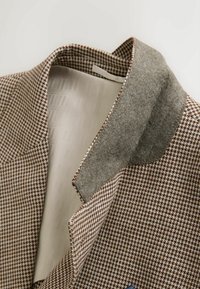 Hnedé a béžové sako so vzorom houndstooth, s šedým filcovým vnútorným golierom a krémovým textúrovaným podšívkou viditeľnou v detailnom záberu.