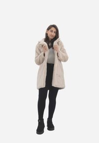 Faux fur jakke i lys beige med hætte, to foran lommer og en skjult lukning. Kombination med sorte leggings og ankelstøvler.