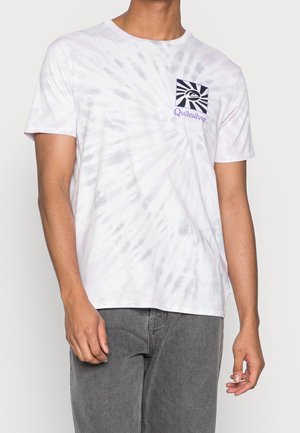Mann trägt ein weiß-graues Batik-Quiksilver-T-Shirt mit schwarzem und lila Logo, kombiniert mit grauer Jeans, steht vor einem schlichten Hintergrund.
