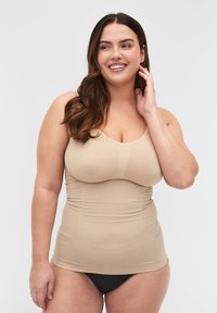 Lächelnde Frau mit langen braunen Haaren, die ein beigefarbenes eng anliegendes Tanktop und schwarze Unterwäsche trägt, posiert vor einem schlichten hellen Hintergrund.