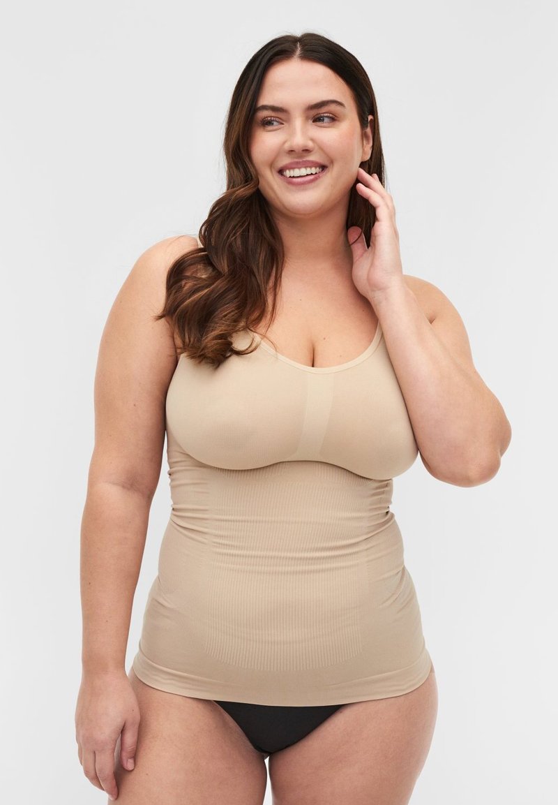 Lächelnde Frau mit langen braunen Haaren, die ein beigefarbenes eng anliegendes Tanktop und schwarze Unterwäsche trägt, posiert vor einem schlichten hellen Hintergrund.
