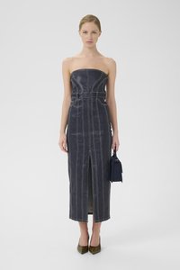 Abito senza spalline in denim scuro con cuciture bianche, vita aderente, spacco laterale e silhouette dritta. La modella tiene una piccola borsa blu.