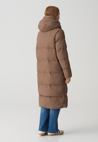 Brauner Daunenmantel mit Kapuze, der ein langes, gestepptes Design aufweist. Kombiniert mit blauen Jeans und weißen Schuhen mit marineblauen Streifen.