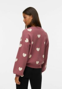 Vrouw met lang donker haar in een mauve trui met witte hartpatronen, staand in een zij-achterhouding tegen een effen achtergrond.