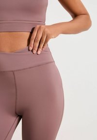 Colanți cu talie înaltă, de culoare mauve, dintr-un material neted și elastic. Prezintă o talie stratificată cu detalii subtile de cusături.