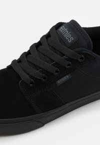 Black sneaker featuring a combination of suede and canvas material, round laces, and a flat rubber sole. Branding on the tongue and side.