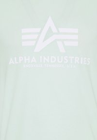 Alpha Industries-logotyp med stiliserat "A" och ränder ovan texten "Alpha Industries Knoxville, Tennessee, USA" på ljus tyg.