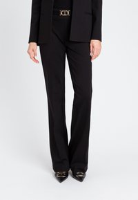 Črne tailored hlače s ravnim krojem, narejene iz gladkega materiala, dopolnjene z elegantno zlatimi sponkami za pas in podolgovatimi črnimi čevlji.