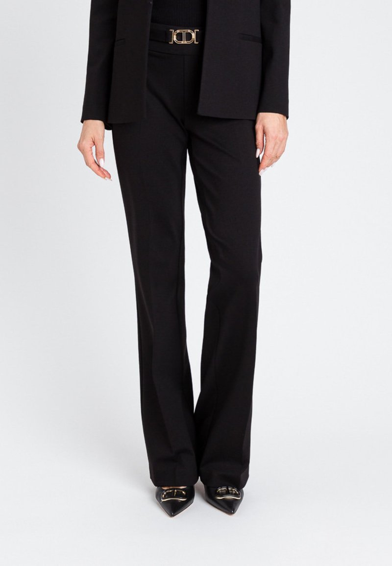 Črne tailored hlače s ravnim krojem, narejene iz gladkega materiala, dopolnjene z elegantno zlatimi sponkami za pas in podolgovatimi črnimi čevlji.