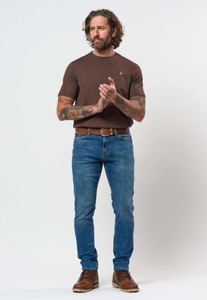 Moški z ramensko dolgimi lasmi in tetovažami, oblečen v rjavo majico, modre kavbojke, rjav pas in usnjene škornje, stoji in drži roke skupaj.