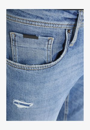 Svijetloplave traperice s leđnim džepom i ušivenom oznakom te istrošenim detaljem na nozi. Klasični dizajn s pet džepova.