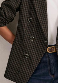 Blazer en pied-de-poule marron avec un design croisé, des boutons noirs et une poche. Porté par-dessus un haut blanc et associé à un jean bleu.