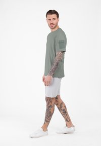 Mężczyzna z tatuażami na rękach w zielonej koszuli z krótkim rękawem, jasnoszarych szortach i białych tenisówkach, patrzący boczkiem na białym tle.