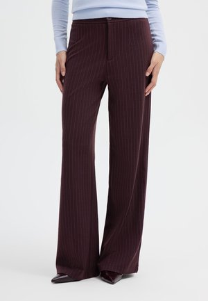 Persona che indossa pantaloni a righe marrone scuro a gamba larga e scarpe nere appuntite, in piedi con le mani appoggiate sulle cosce.