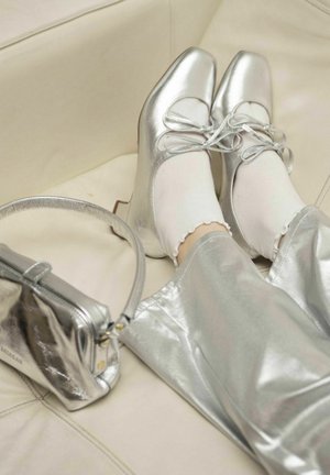 Scarpe con tacco in metallo argento con dettagli in pizzo, abbinate a calze bianche e pantaloni argento lucidi. Una borsa a tracolla metallizzata coordinata è visibile.