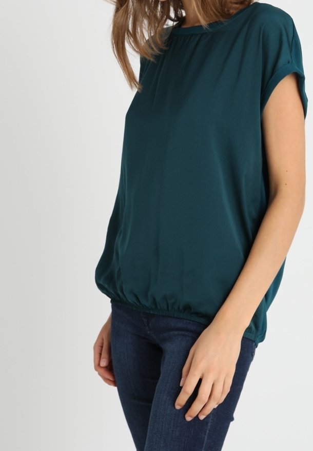 Femme portant un chemisier à manches courtes bleu sarcelle avec un ourlet élastique, associé à un jean skinny bleu foncé, se tenant devant un fond blanc uni.