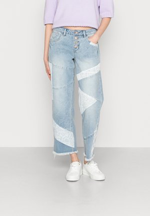 Jeans de denim azul claro com design em patchwork, apresentando acentos brancos em padrão, fecho frontal com cinco botões e bainha cropped com efeito desfiado.