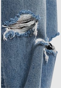 Vääristatud teksad keskmiselt sinises denimis, millel on rebenditega augud. Omab sirge sääre disaini ja tekstuurset kangast koos nähtavate õmblusdetailidega.