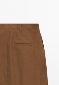 Section de pantalon marron montrant la ceinture, la boucle de ceinture et une poche arrière passepoilée simple sur fond blanc uni.