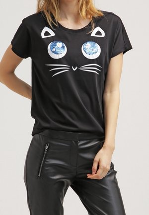 Kobieta nosząca czarny t-shirt z wzorem kota z niebieskimi cekinowymi oczami, w połączeniu z czarnymi skórzanymi spodniami z kieszenią na zamek.