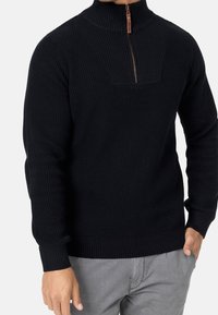 Homme portant un pull en maille texturée noir avec une fermeture éclair quart de zip et un tirant marron, un pantalon gris, main dans la poche, sur un fond blanc.