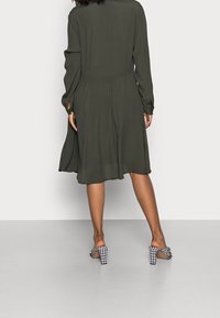 Kobieta ubrana w ciemnozieloną midi sukienkę z długimi rękawami i czarno-białe sznurowane sandały na wysokim obcasie, stojąca na szarym podłożu.