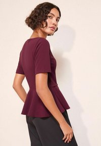 Burgundská peplum blůza s krátkými rukávy, se přiléhavým tělem a rozšířeným lemem. Hladká textura; kombinováno s tmavými, upravenými kalhotami.