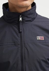 Marinblå vindjacka med hög krage, framdragkedja och en liten broderad logga med röda och blå element på bröstet.