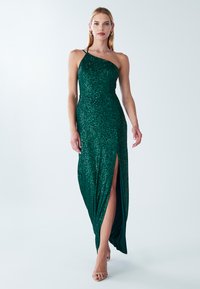 Žalia vakarinė suknelė su blizgančiomis akutėmis, asimetriniu iškirptės dizainu, šoniniu plyšiu ir prigludusiu siluetu. Lygi tekstūra ir blizgi apdaila.