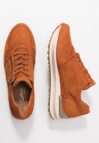 Ett par brända orange mockasneakers med snören, sidoflik, och vita gummisulor, visade från ovan och sidovinklar på vit bakgrund.