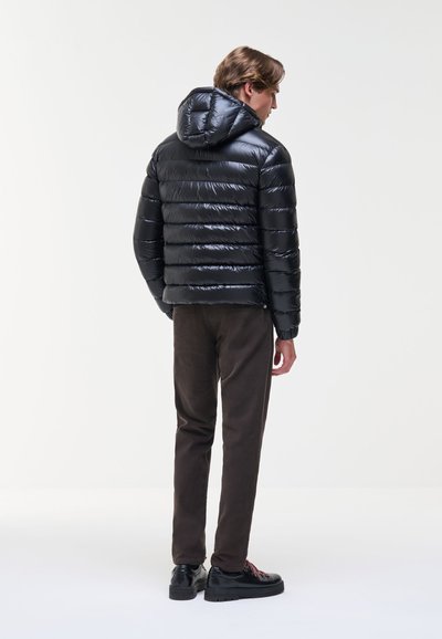 Homme debout de dos portant une veste matelassée noire brillante avec capuche, un pantalon foncé et des chaussures noires sur un fond clair uni.