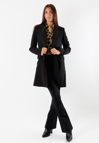 Cappotto nero con colletto a reversal e due tasche frontali, indossato sopra un top in stampa leopardo dorata e pantaloni neri a palazzo. Stivali neri con tacco completano l'outfit.