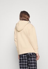 Sweat à capuche beige avec une grande capuche, des poignets et un ourlet côtelés. Associé à un pantalon à carreaux noir et blanc. Texture de tissu lisse.