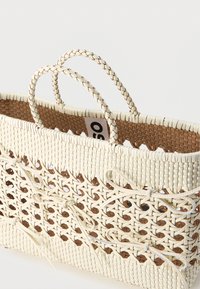 Λευκή υφαντή τσάντα χειρός με πλεγμένες λαβές και διακοσμητικούς δερμάτινους φιόγκους σε ταμπλέ φόντο.