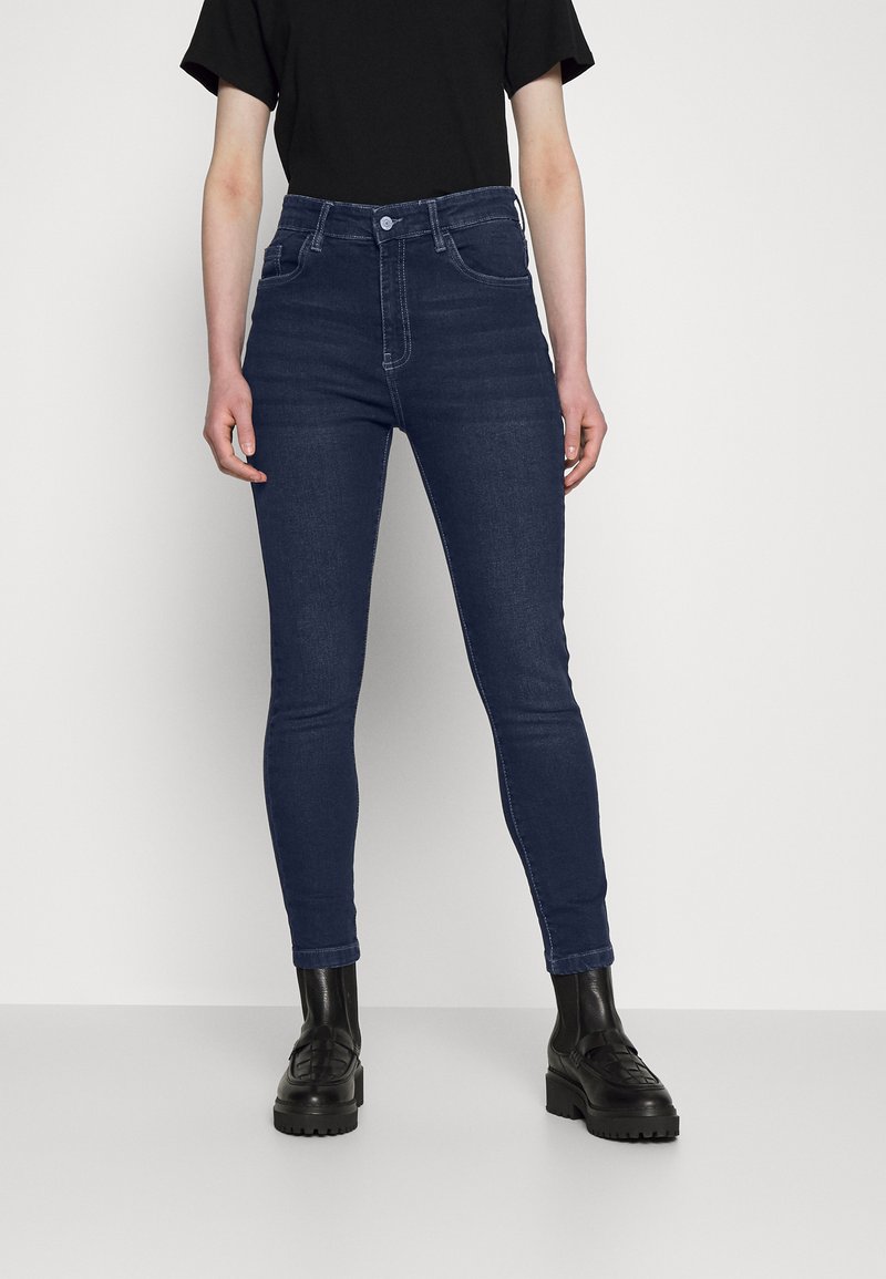 Jeans de cintura alta azul escuro com um corte slim e ligeira elasticidade, apresentando bolsos traseiros e uma textura de denim suave. Combinados com botas pretas.