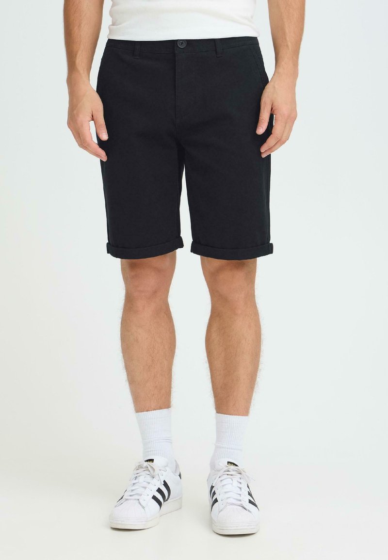 Shorts en coton noirs avec revers, dotés d'une fermeture à bouton et de poches latérales, portés avec des chaussettes blanches et des baskets noires à rayures.