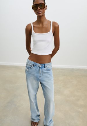 Top corto bianco senza maniche, jeans larghi azzurri con dettagli consumati e occhiali da sole marroni oversize. Design minimalista con vestibilità aderente.