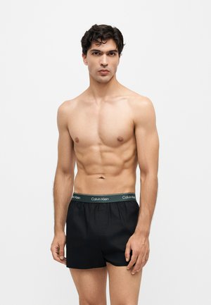 Tânăr fără cămașă cu păr închis la culoare, purtând boxeri negri Calvin Klein pe un fundal simplu și deschis la culoare.